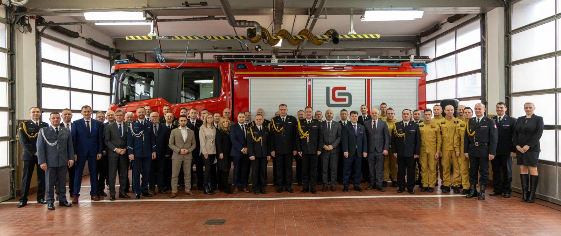 Grupowe zdjęcie kilkudziesięciu osób stojących w hali strażackiej przed czerwonym wozem strażackim. Uczestnicy są ubrani w mundury służbowe, garnitury oraz specjalistyczne kombinezony. Po bokach duże bramy garażowe.