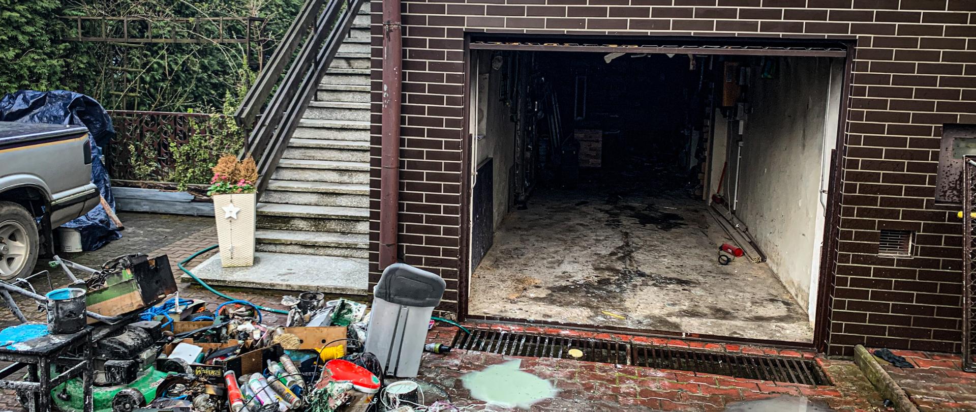W środkowej części zdjęcia jest garaż otwarty który znajduje się w budynku mieszkalnym, po lewej stronie znajduje się spalona kosiarka oraz akcesoria użytku domowego, po lewej stronie znajdują się schody z wejściem do domu oraz jest tył pojazdu terenowego