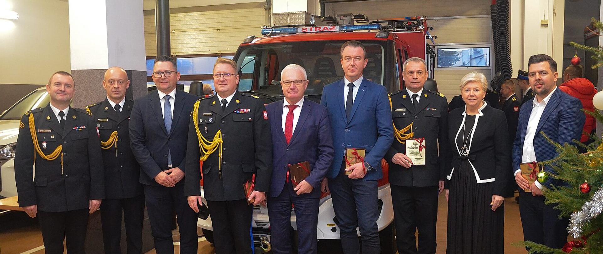 komendanci oraz politycy i przedstawiciele samorządu pozują do zdjęcia w tle stoi lekki samochód strażacki iveco ozdobiony biało-czerwonymi wstęgami
