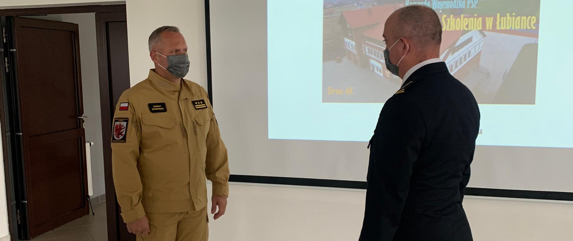 Na zdjęciu widać dwóch strażaków stojących naprzeciw siebie w tle slajd prezentacji na ekranie rzutnika.