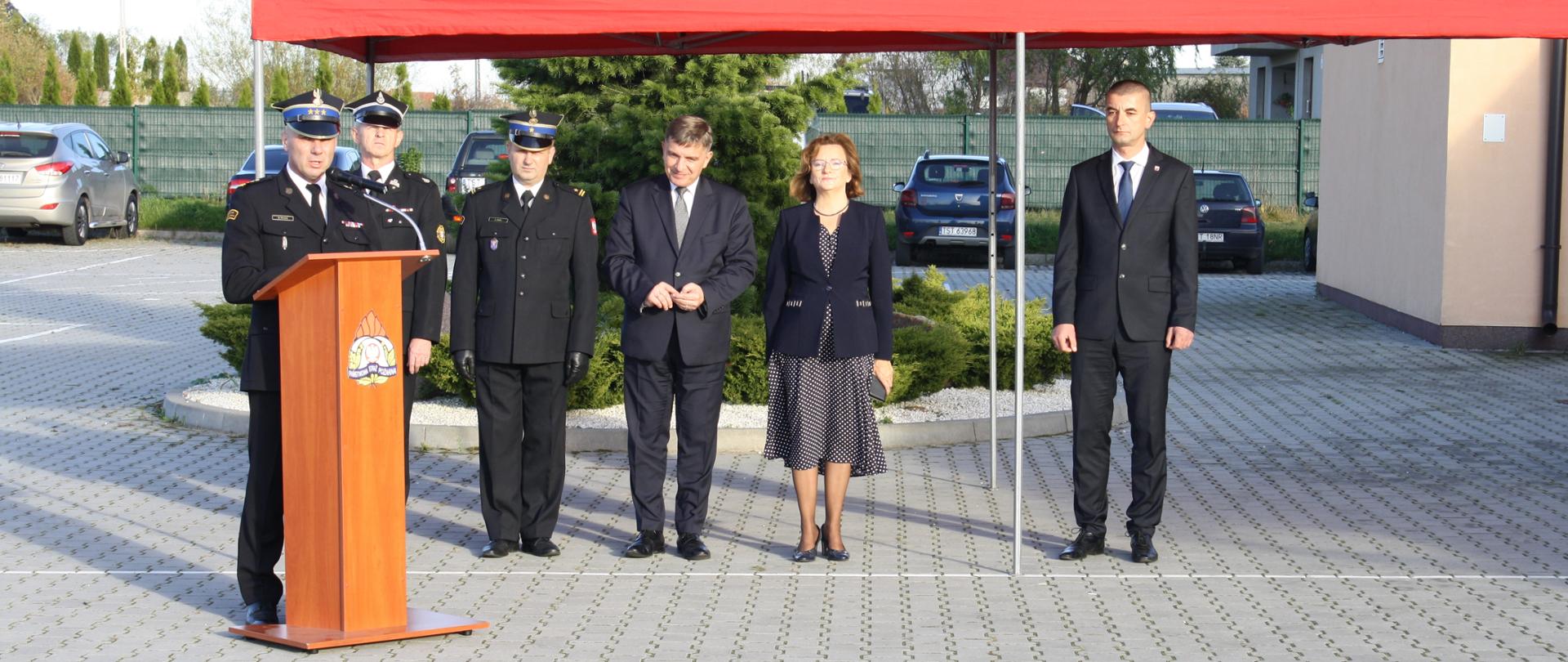 Na pierwszym planie zaproszeni goście w odświętnych strojach stoją pod czerwonym namiotem. Zastępca Świętokrzyskiego Komendanta Wojewódzkiego PSP w mundurze galowym przemawia z mównicy 