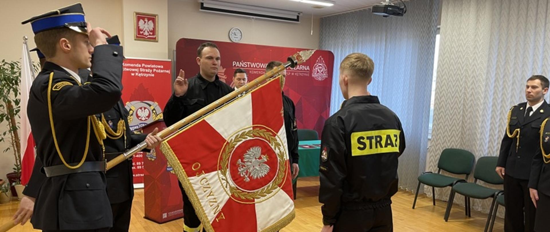Strażacy ślubują przed sztandarem trzymając dwa palce (środkowy i wskazujący) prawej dłoni w górze. Sztandar jest biało-czerwony, posiada złote litery oraz złote boki. Na jego środku widnieje godło Polski.
