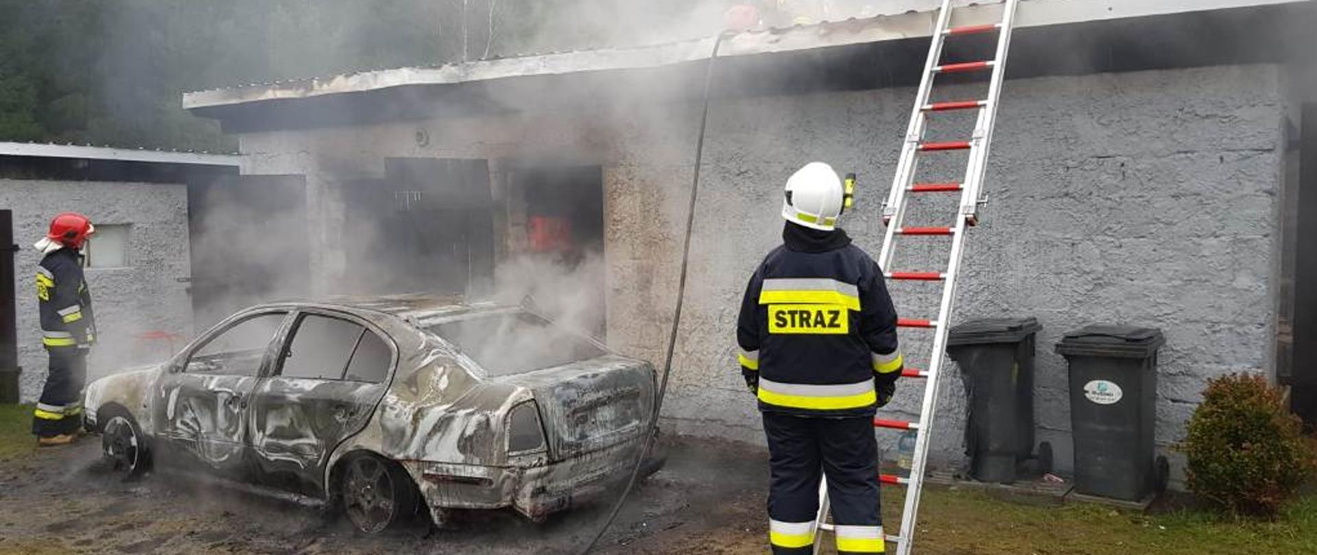 Na tle budynku widać dwóch strażaków stojących koło spalonego samochodu osobowego. Strażak stojący z prawej strony zabezpiecza drabinę opartą o dach budynku. 