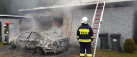 Dwóch strażaków stoi koło spalonego samochodu, z którego wnętrza nadal unosi się dym. Jeden z nich zabezpiecza drabinę opartą o zagrożony budynek.