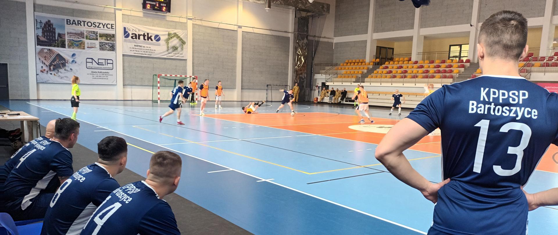 Na pierwszym planie zawodnicy na ławce rezerwowej oraz stojący zawodnik z napisem na plecach KP PSP Bartoszyce, na drugim planie zawodnicy rozgrywający mecz