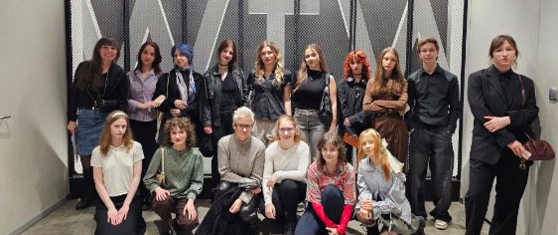 Na zdjęciu wiele postaci młodych ludzi. Dziesięć osób stoi w rzędzie, przed nimi jeszcze sześć osób przyklęknęło na podłodze. W tle szare ściany pomieszczenia teatru i litery WTW - Wrocławski Teatr Współczesny. 