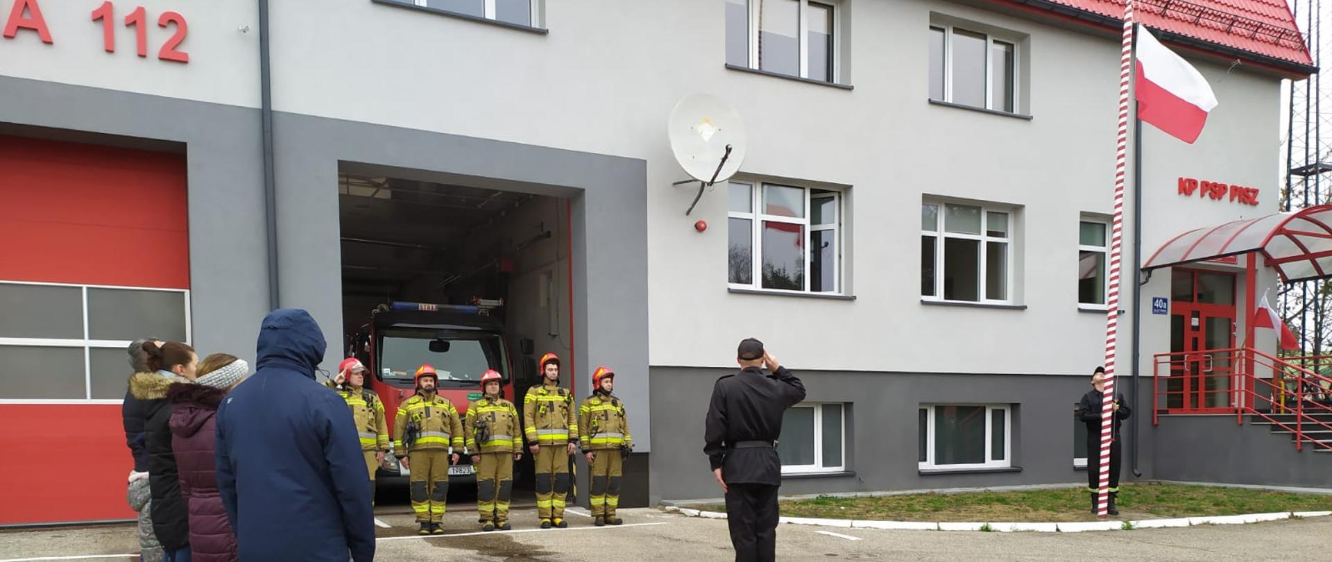 Strażacy stoją w szeregu przed Komendą podnosząc flagę na maszt.