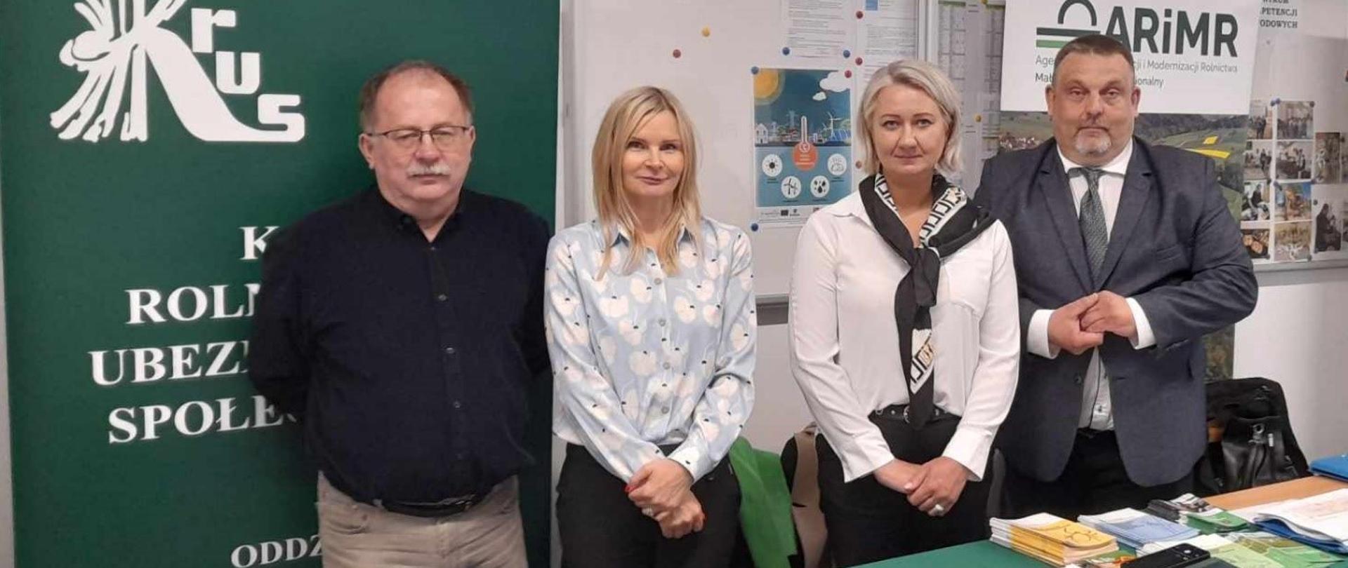 Dwie kobiety i dwóch mężczyzn pozują do zdjęcia za stołem przykrytym zielonym suknem z rozłożonymi ulotkami i broszurami KRUS, za nimi stoi zielony rollup z logo KRUS, na ścianie wiszą gabloty szkolne.