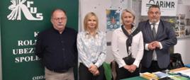 Dwie kobiety i dwóch mężczyzn pozują do zdjęcia za stołem przykrytym zielonym suknem z rozłożonymi ulotkami i broszurami KRUS, za nimi stoi zielony rollup z logo KRUS, na ścianie wiszą gabloty szkolne.