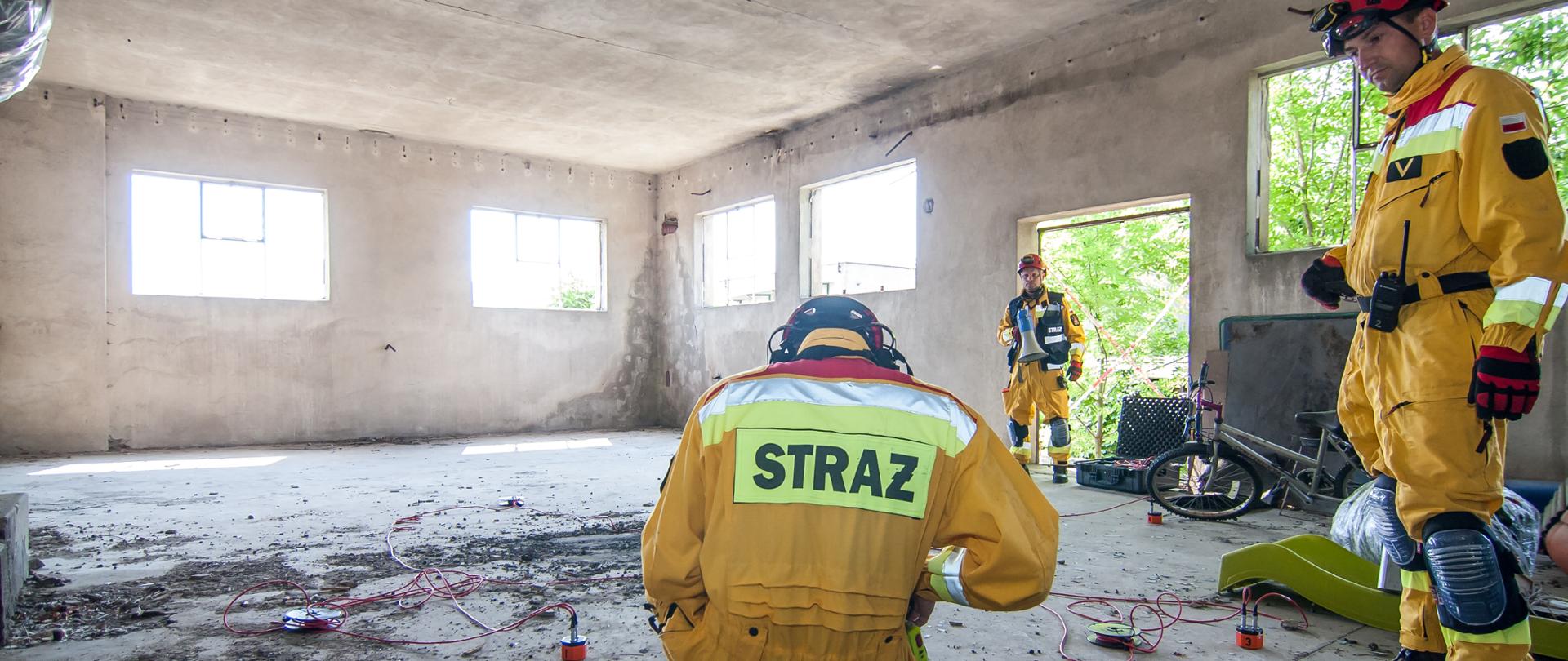 Zdjęcie przedstawia trzech strażaków w kombinezonach koloru żółtego z czerwonymi kaskami na głowie w pustym pomieszczeniu. Na podłodze rozstawione czujniki geofonu spięte ze sobą czerwonymi kablami. Jeden ze strażaków kuca na środku kadru, pozostali strażacy stoją po jego prawej stronie. W oddali leży rower oraz część plastikowej zjeżdżalni dla dzieci.