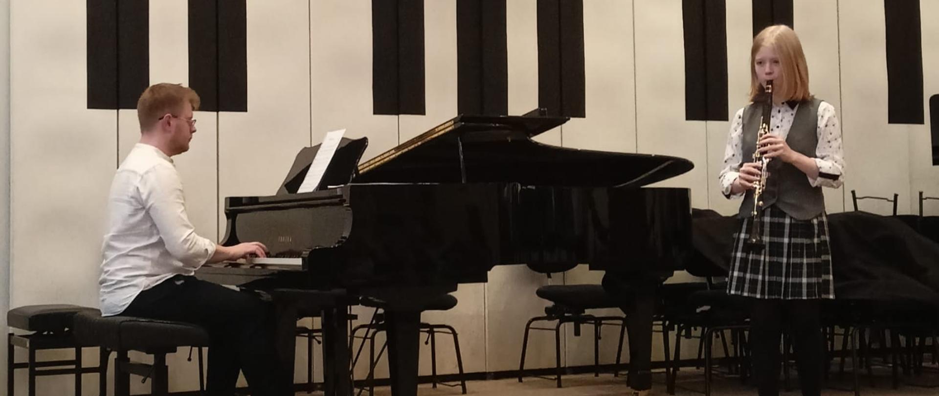 Dziewczynka stojąc po prawej stronie na scenie auli PSM gra na klarnecie, tuż za nią po lewej stronie mężczyzna gra na fortepianie.