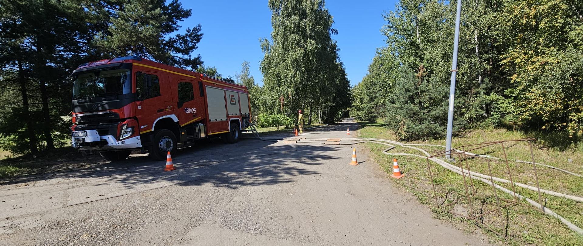 Samochód gaśniczy Państwowej Straży Pożarnej ustawiony przy leśnej drodze, obok rozłożone węże tłoczne i pachołki drogowe. W głębi widoczny strażak w hełmie nadzorujący działania.