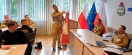 Zdjęcie zrobione w sali konferencyjnej podczas szkolenia członków pocztów sztandarowych. Strażak prowadzi wykład, a uczestnicy siedzą przy stołach konferencyjnych.