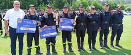 Zdjęcie przedstawia 8 osób stojących w rzędzie. Od lewej strony stoi mężczyzna ubrany w białą koszulę i granatowe spodnie, następnie 4 strażaków, 1 strażaczka i 2 strażaków. Wszyscy ubrani w stroje koszarowe. Trzech strażaków od lewej trzyma tabliczki za napisaną kwotą nagrody za I-III miejsce oraz puchary.
