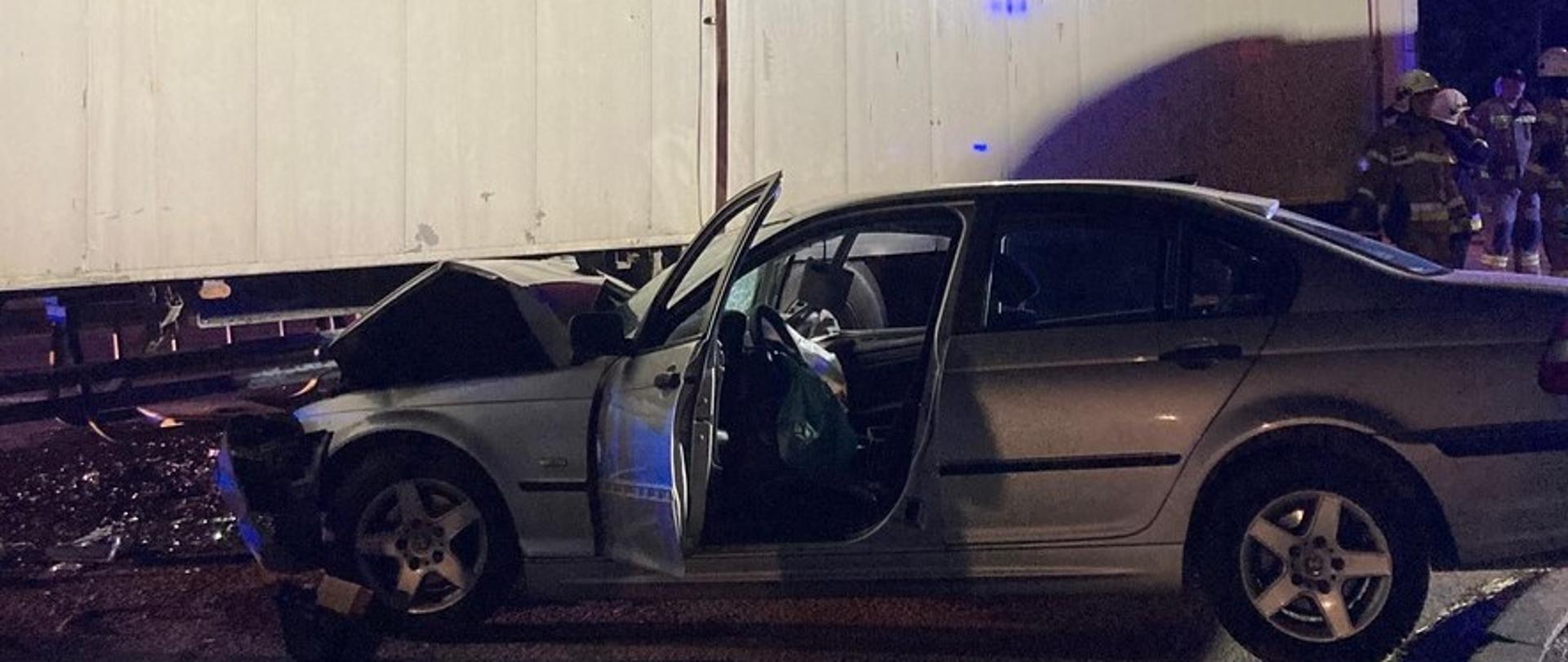 Od lewej strony widać ciągnącą się naczepę samochodu ciężarowego. Pod naczepę lekko wsunięty stoi samochód osobowy ze zniszczonym przodem. W głębi stoi grupa strażaków.