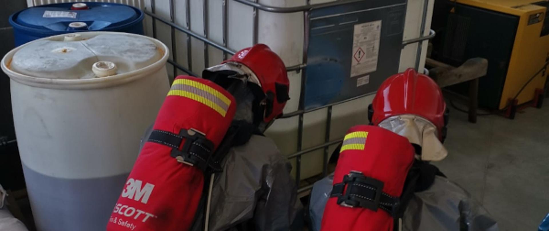 Dwóch strażaków w kombinezonach specjalnych w kolorze szarym, w czerwonych butach, z czerwonymi butlami z tlenem na plecach oraz czerwonych hełmach klękają przez pojemnikiem w części wypełnionym substancją płynną, obok widać dwie beczki - niebieską oraz białą wypełnioną do połowy, w tle okna budynku 