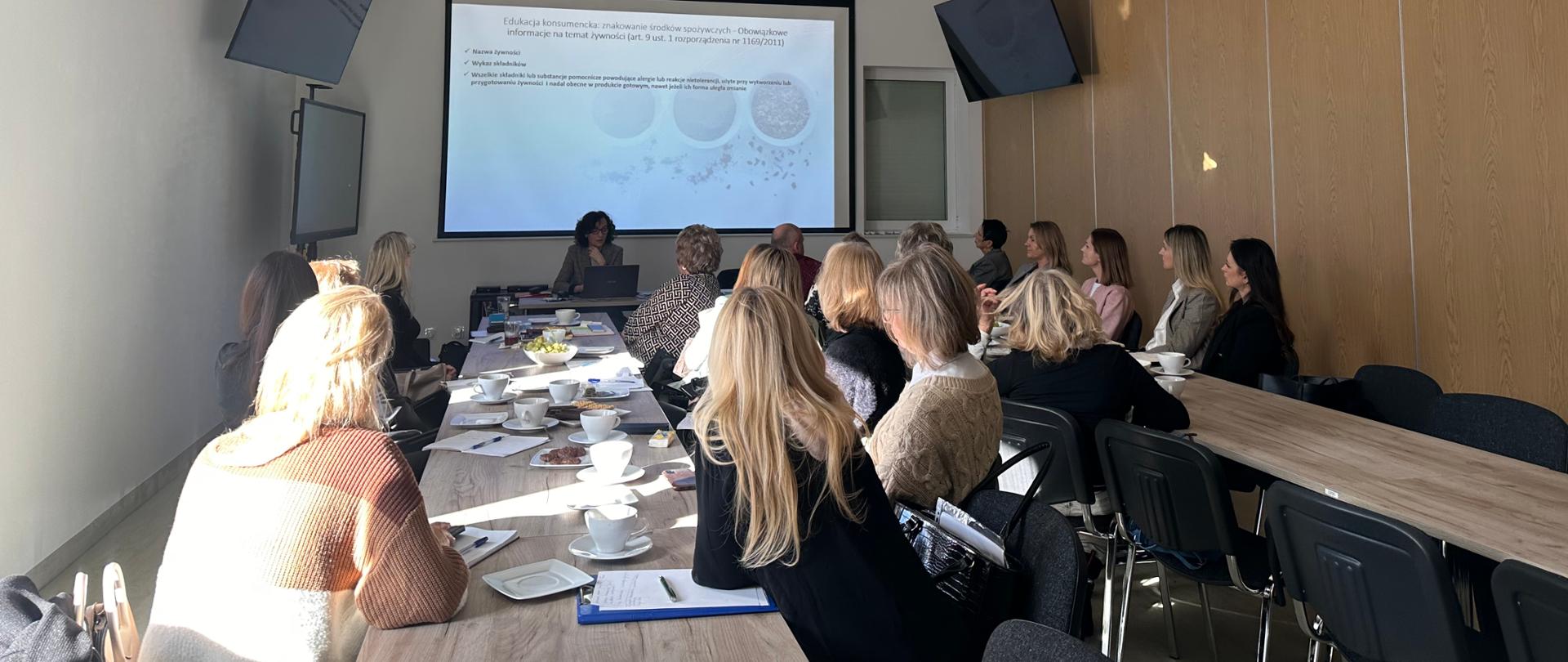 Na zdjęciu widzimy wyświetlaną prezentację, osobę która przeprowadza szkolenie oraz uczestników szkolenia.