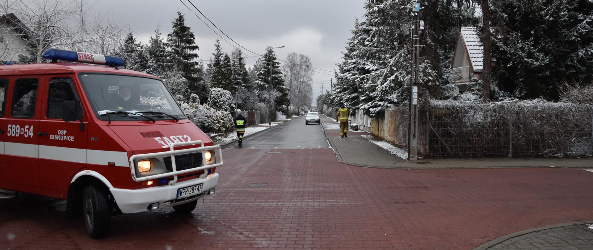 Zdjęcie przedstawia proces dystrybucji ulotek przez członków ochotniczych straży pożarnej. Na tle ulicy widać samochód pożarniczy oraz dwoje strażaków roznoszących ulotki. 