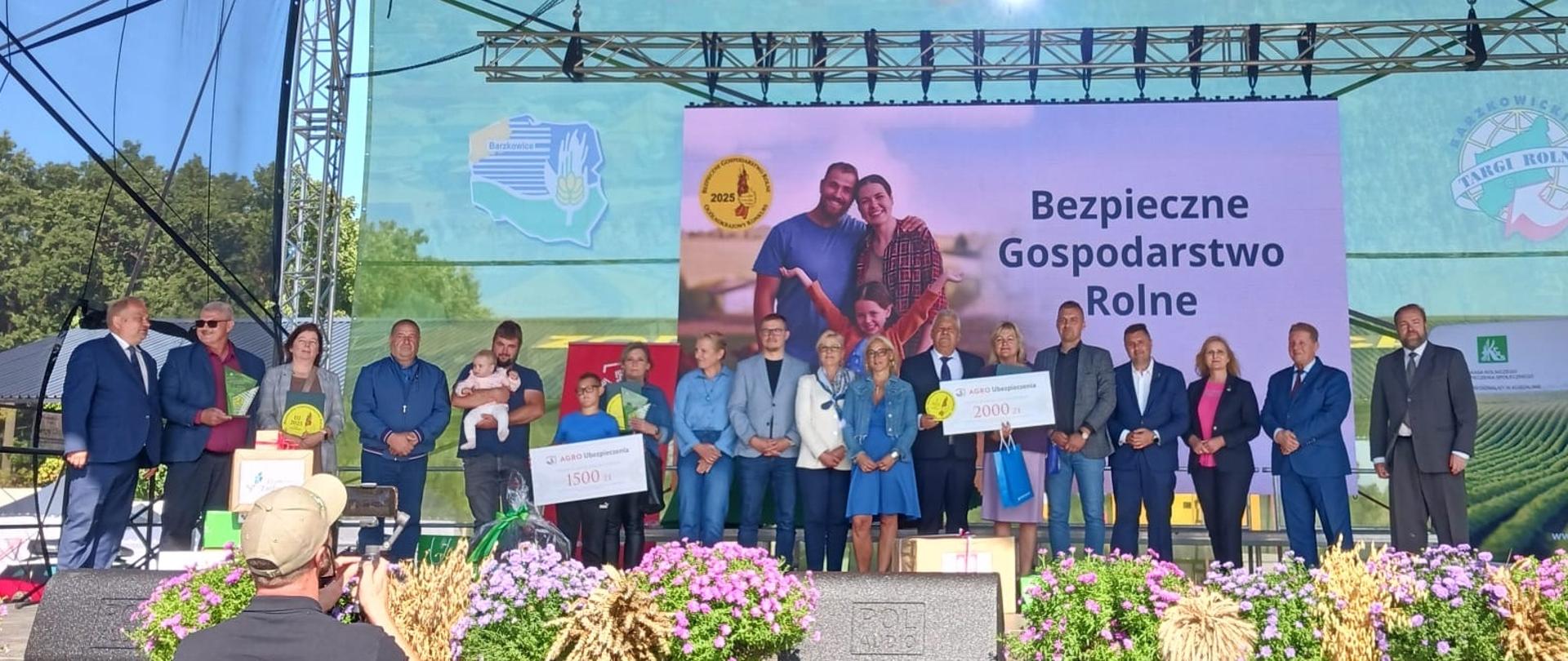 Na scenie stoi duża grupa osób dorosłych i kilkoro dzieci. Dwie osoby trzymają w rękach makiety czeków z kwotą nagrody pieniężnej, żółte okrągłe dyplomy z logo BGR oraz torby z nagrodami rzeczowymi.