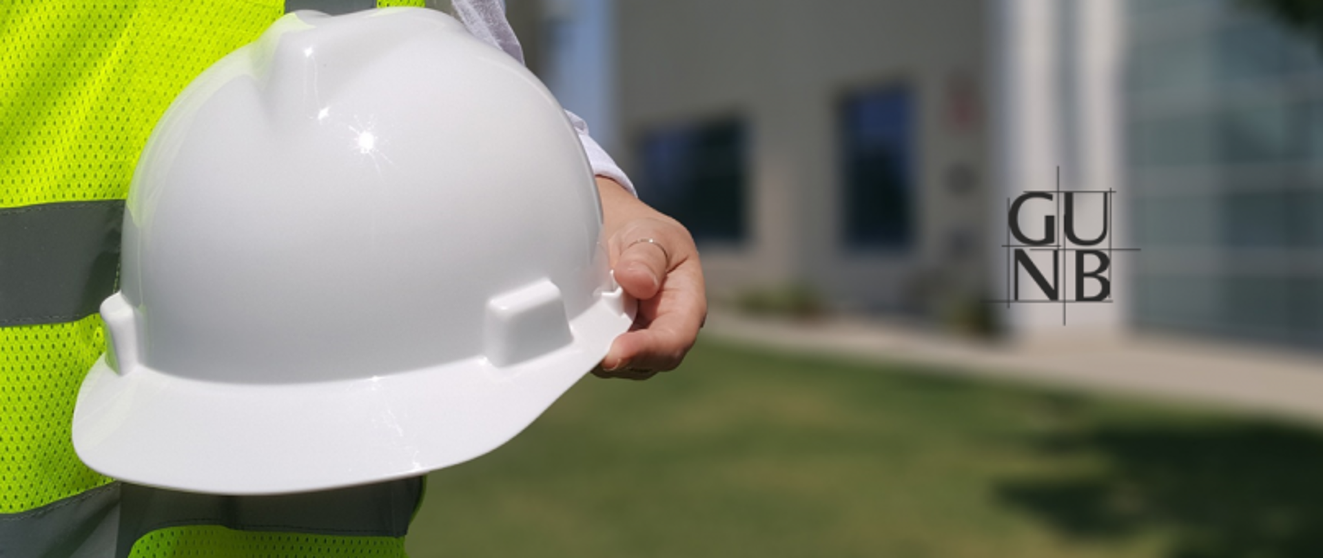 Osoba w kamizelce odblaskowej trzymająca biały kask; w tle budynek i zieleń, po prawej stronie logo GUNB.