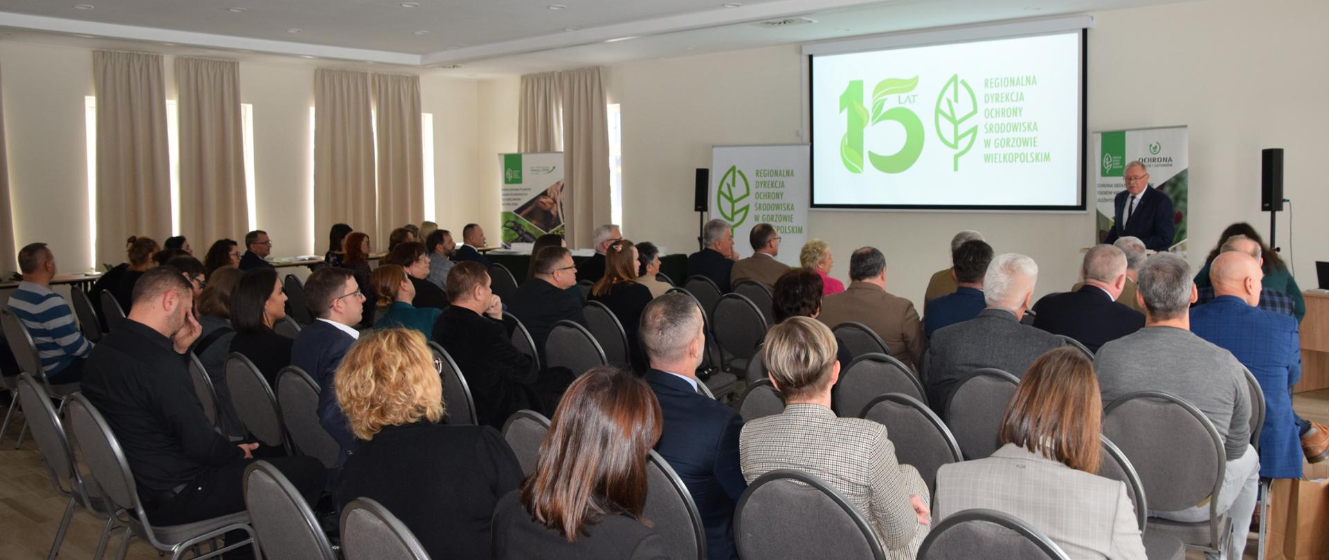 W dużej sali na krzesłach siedzi grupa kilkudziesięciu osób odwróconych plecami. Na ścianie przed na drugim planie wisi duży ekran z wyświetloną prezentacją.