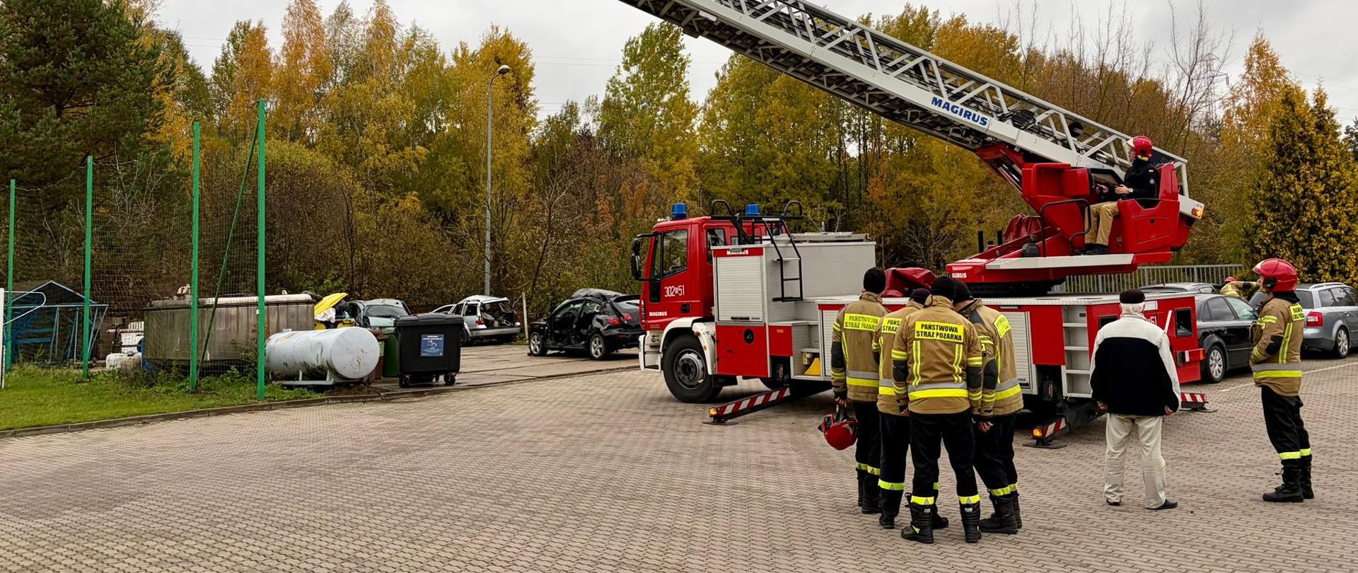 Szkolenie doskonalące dla kierowców samochodów z drabiną mechaniczną - strażacy stoją na placu obok podnośnika w trakcie szkolenia, w tle zaparkowane samochody i jesienne drzewa