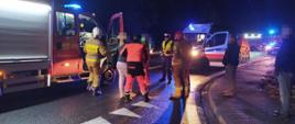 Zderzenie dwóch aut. Droga wojewódzka nr 309 w okolicach Żylic. Zespół ratownictwa medycznego prowadzi poszkodowaną do ambulansu. Na jezdni stoi karetka pogotowia i wozy straży pożarnej., a także policjant i strażacy.