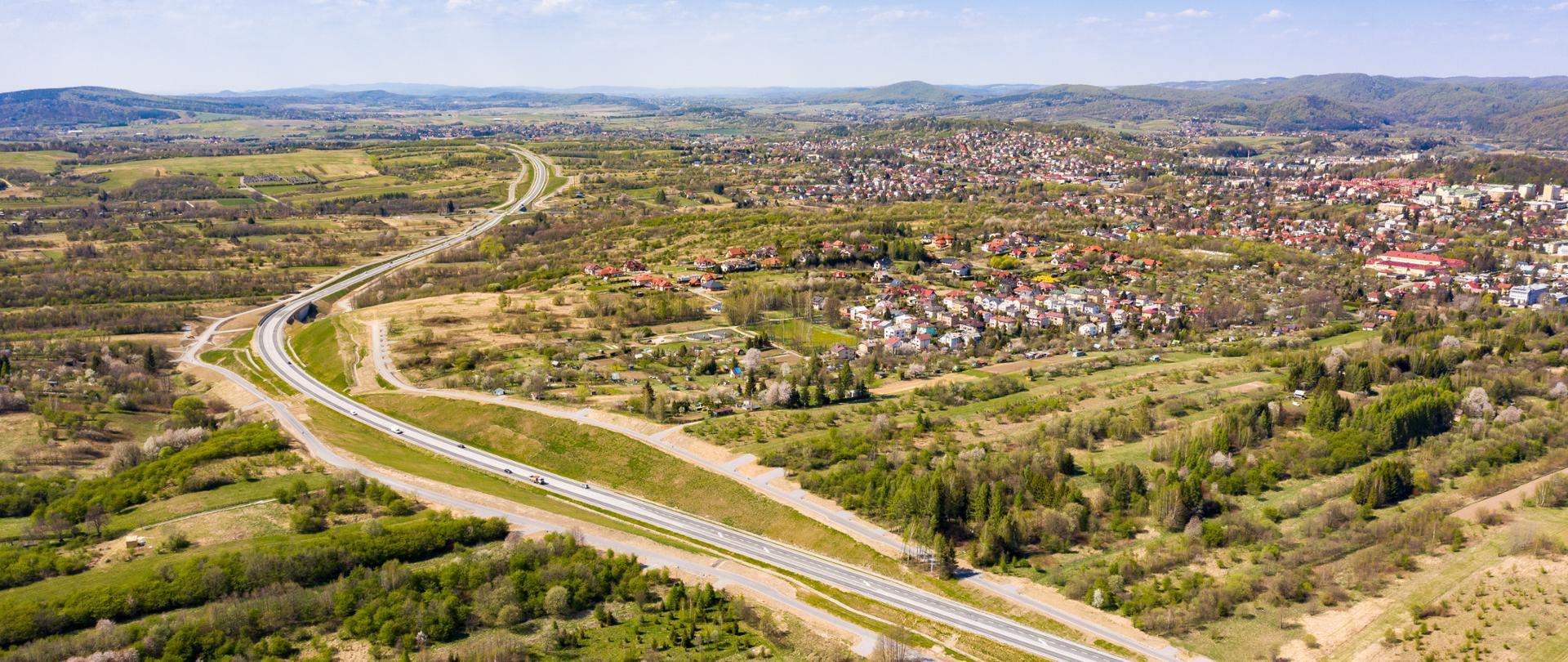 Widok z lotu ptaka na przebieg obwodnicy Sanoka. Lato, świeci słońce, po obu stronach drogi widać tereny zielone. Po drodze jadą auta. Droga omija z lewej strony zabudowania. 