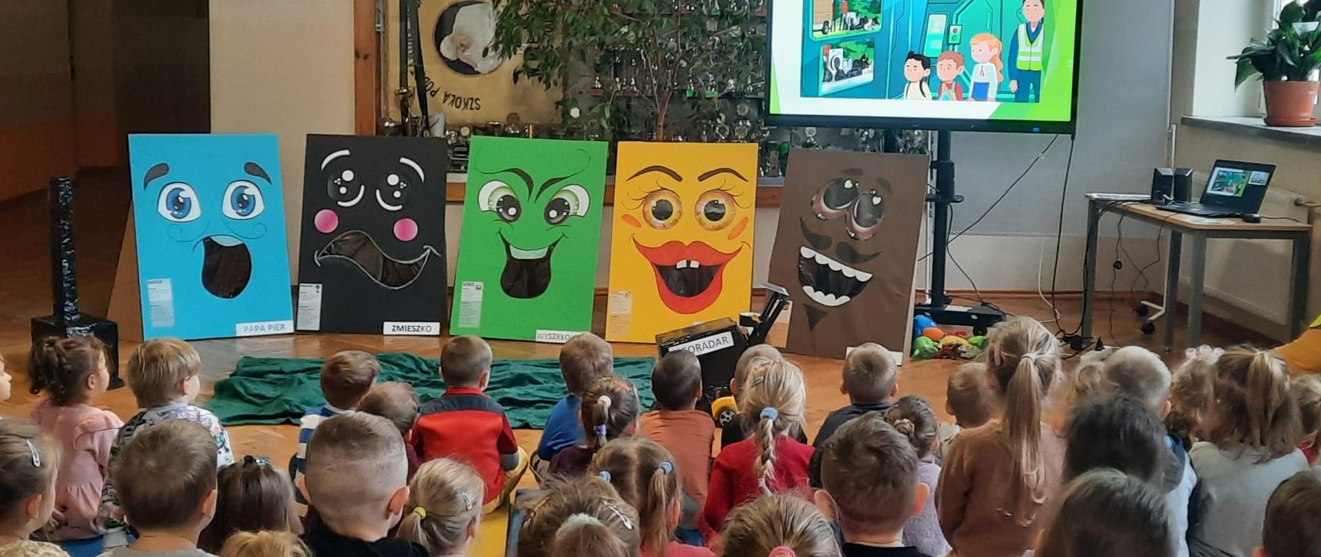 1. Dzieci siedzące na podłodze oglądają film kreskówkowy związany z ochroną środowiska. Obok telewizora postawione są cztery kartonowe postacie. Niebieski karton o nazwie Papa Pier, czarny o nazwie Zmieszko, zielony o nazwie Wyszkłolony, żółty o nazwie Plastika Metalika, brązowy o nazwie El Bio. Wszystkie postacie kartonowe mają duże oczy i robią śmieszne miny. Przed dziećmi leży też zielony materiał, który przykrywa odpady i obok stoi czarny Georadar.