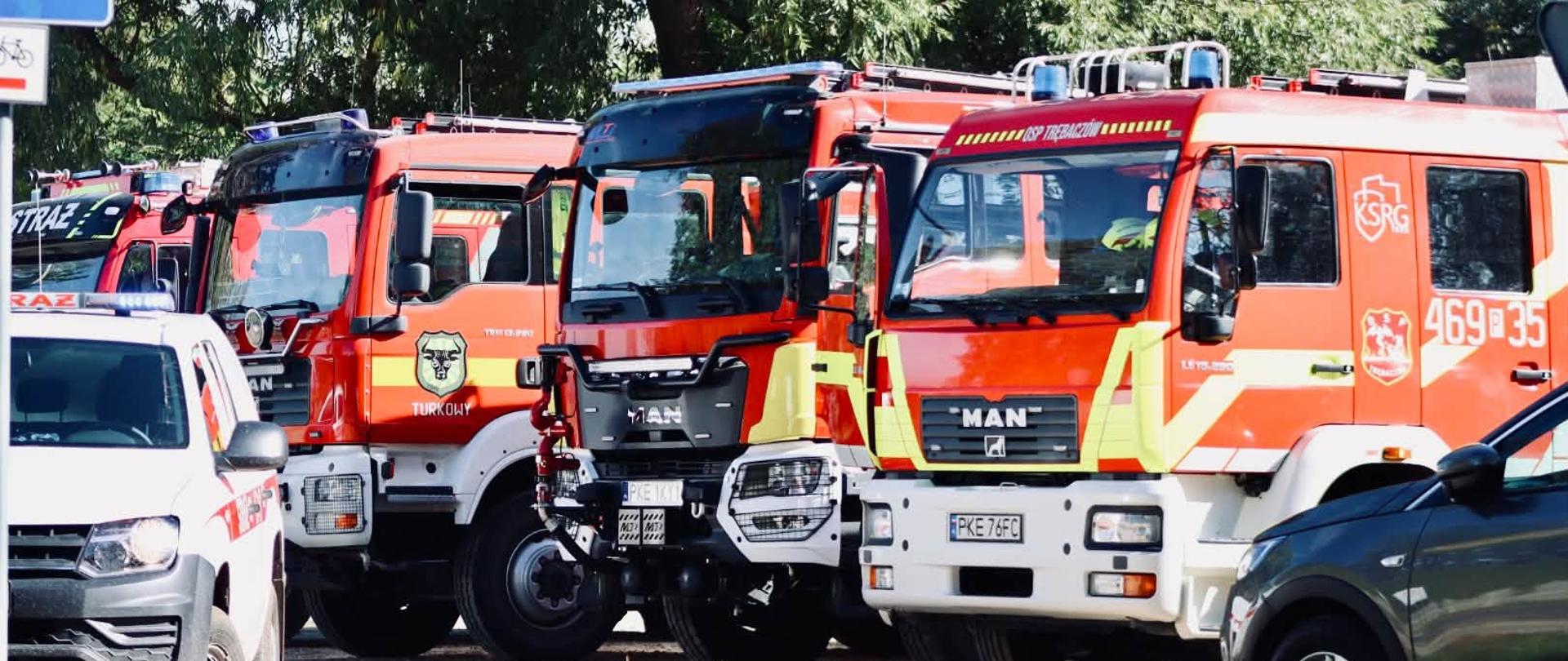 Czerwone wozy strażackie marki MAN należące do Ochotniczej Straży Pożarnej zaparkowane w rzędzie w otoczeniu zieleni. Na pierwszym planie widoczne są fragmenty pojazdów cywilnych.