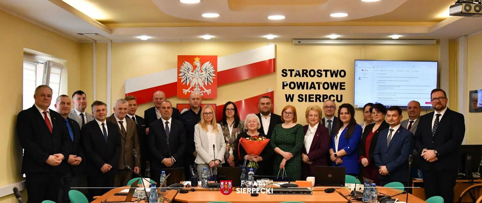 Grupa kilkunastu osób w formalnych strojach stoi w sali konferencyjnej przed stołem, pod godłem Polski i napisem „Starostwo Powiatowe w Sierpcu”; w centrum kobieta trzyma bukiet kwiatów.