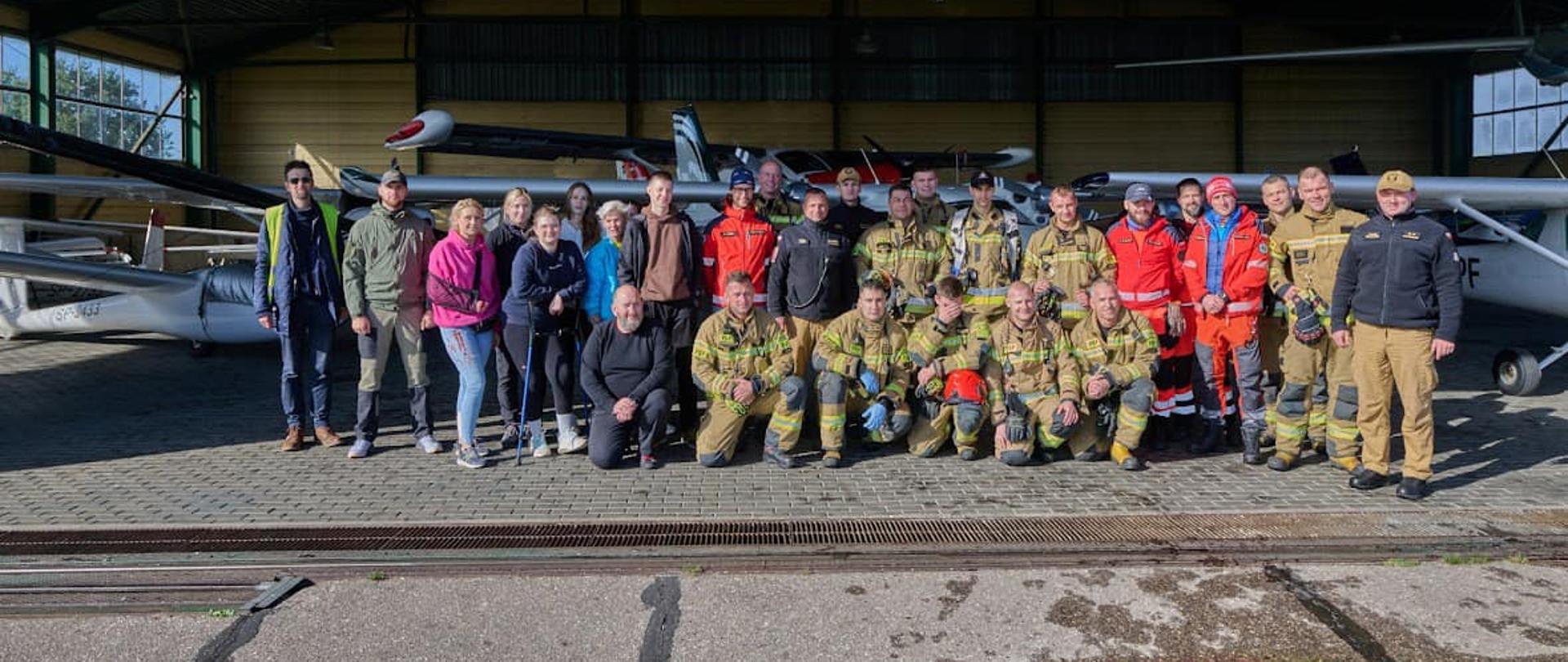 Na zdjęciu znajdują się osoby uczestniczące w ćwiczeniach na elbląskim lotnisku. Są to strażacy, policjanci oraz ratownicy medyczni stanowiący załogi ZRM. Osoby znajdują się w hangarze lotniska, na tle samolotu,