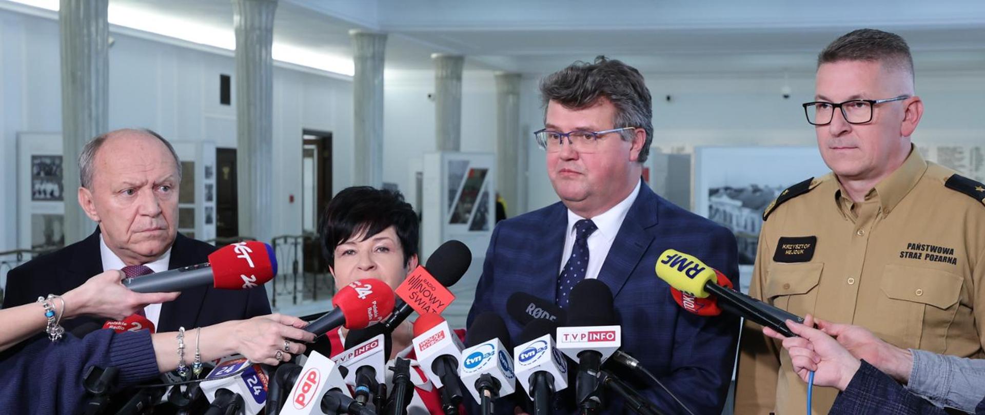 Osoby cywilne stoją oraz funkcjonariusz Państwowej Straży Pożarnej stoją obok siebie przed mikrofonami.