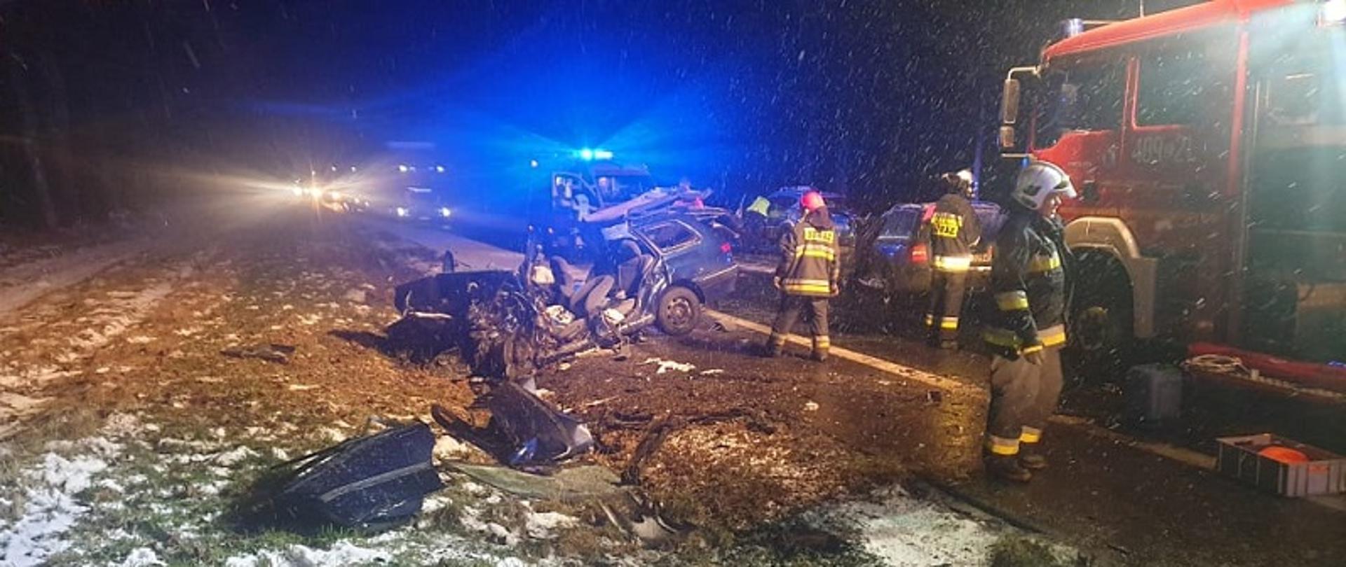Zdjęcie przedstawia miejsce wypadku wykonane nocą. Na pierwszym planie znajduje się wrak pojazdu widoczny od strony kierowcy. Samochód przodem skierowany w stronę pobocza. Po prawej stronie wraku znajduje się samochód strażacki i trzech strażaków w ubraniach specjalnych. Miejsce wypadku oświetlone sztucznie przez pojazdy pożarnicze, widoczne opady śniegu i pobocze drogi z pokrywą śnieżną