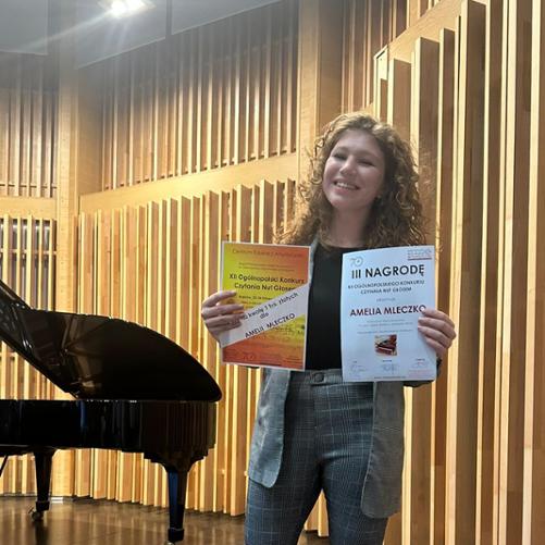 Elegancko ubrana nastolatka stoi przed fortepianem i trzyma w rękach dwa dyplomy.