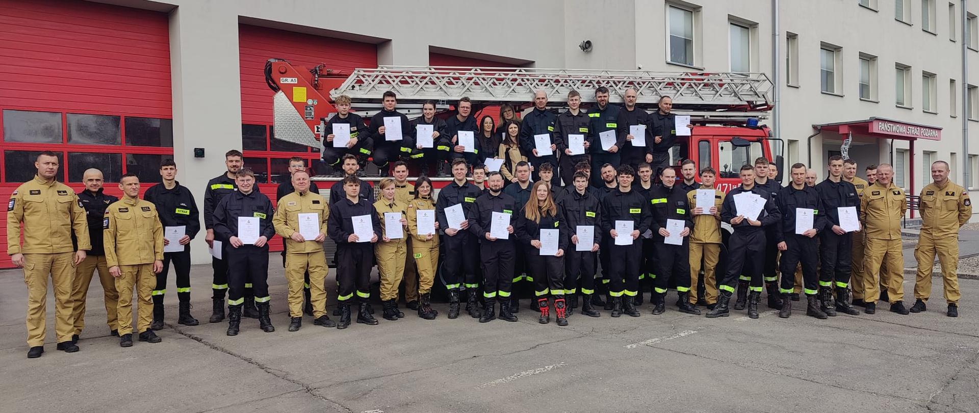 Grupa strażaków w ubraniach koszarowych koloru czarnego i piaskowego trzymająca zaświadczenia ukończenia szkolenia pozuje na tle samochodu bojowego typu drabina i budynku komendy powiatowej.