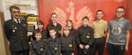 Grupa dzieci i młodzieży w strojach strażackich pozuje do zdjęcia wraz z umundurowanym strażakiem w sali wydarzenia. Uczestnicy stoją w jednym rzędzie na tle czerwonej ścianki z emblematem straży pożarnej oraz baneru organizacyjnego. Niektóre dzieci mają na sobie mundury z odblaskowymi elementami, a atmosfera jest oficjalna, ale jednocześnie przyjazna.
