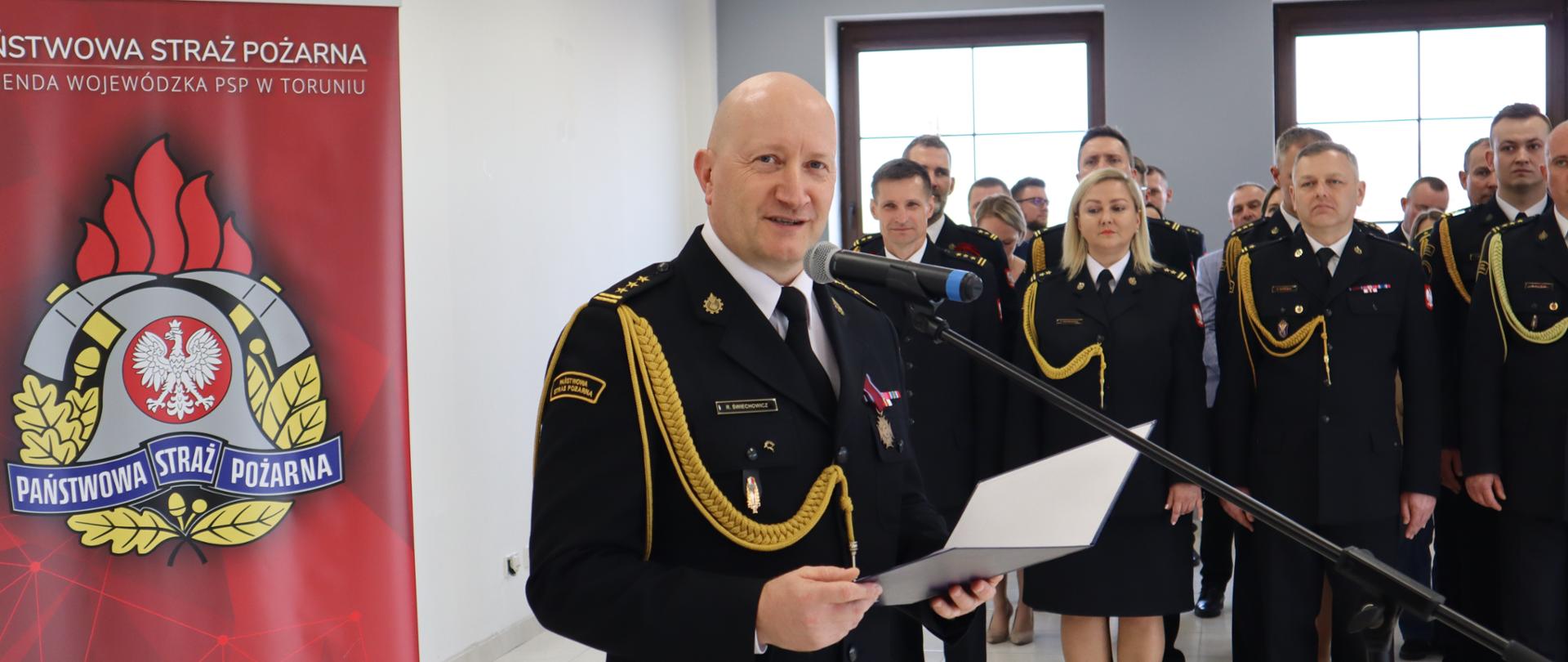 Strażak w mundurze galowym stoi przy mikrofonie. W ręku trzyma dokument. W tle pododdział strażaków i z lewej strony baner firmowy.