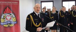 Strażak w mundurze galowym stoi przy mikrofonie. W ręku trzyma dokument. W tle pododdział strażaków i z lewej strony baner firmowy.