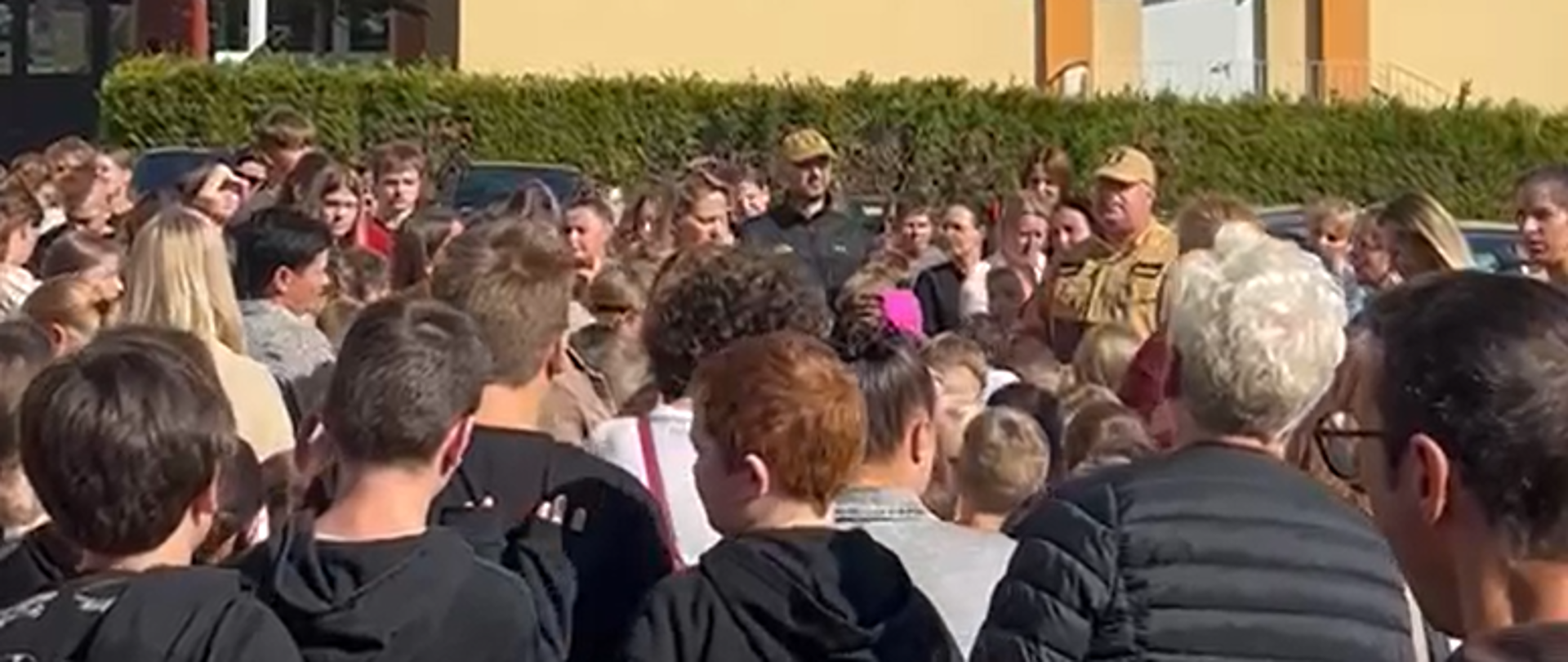 Zdjęcie przedstawia zgromadzenie uczniów, nauczycieli oraz strażaków Państwowej Straży Pożarnej na zewnątrz budynku szkolnego podczas praktycznych ćwiczeń z zakresu ewakuacji.
**Główne elementy na zdjęciu:**
* **Uczestnicy ćwiczeń:** Zgromadzona grupa składa się z młodzieży w różnym wieku, dorosłych (prawdopodobnie kadry pedagogicznej) oraz strażaków w piaskowych/ciemnych mundurach służbowych. Wszystkie osoby stoją blisko siebie, uwagę kierując w stronę prowadzących omówienie.
* **Strażacy Państwowej Straży Pożarnej:** W środkowej części tłumu widoczni są strażacy w czapkach z daszkiem i charakterystycznych mundurach PSP, którzy prawdopodobnie podsumowują przebieg ewakuacji i przekazują uwagi dotyczące bezpieczeństwa.
* **Otoczenie:** Akcja odbywa się na placu przed nowoczesnym, żółto-piaskowym kompleksem budynków szkolnych. W tle widać wysokie drzewo iglaste, niski żywopłot oraz fragmenty zaparkowanych pojazdów. Pogoda jest słoneczna, a niebo jasne i bezchmurne.