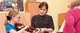 Nauczycielka Natalia Mierzwa, która poprawia uczennicy ułożenie rąk na skrzypcach , po prawej stronie stoi przyglądająca się P. Danuta Wolnomiejska