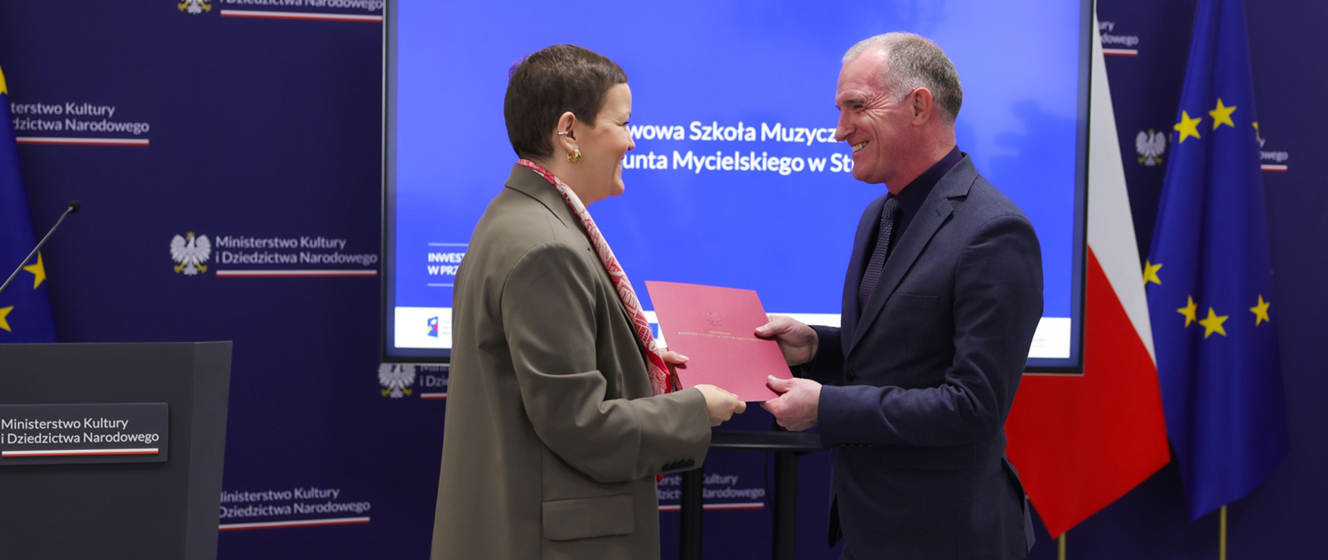 Baner w ciemnoniebieskiej kolorystyce z powtarzającym się napisem „Ministerstwo Kultury i Dziedzictwa Narodowego” oraz godłem Polski. Na środku znajduje się duży ekran z niebieskim tłem i białym napisem informującym o państwowej szkole muzycznej oraz inwestycji. Po prawej stronie widoczne są stojące flagi Polski i Unii Europejskiej. Na tle baneru stoją dwie osoby w formalnych strojach, przekazujące sobie czerwony dokument.