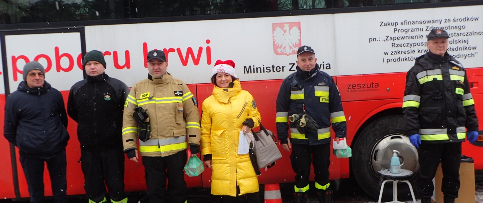 Strażacy OSP i PSP w mundurach oraz cywile stoją przed krwiobusemw. W środku kobieta w żółtej kurtce i czapce mikołaja. W tle krwiobus.