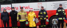Strażacy OSP i PSP w mundurach oraz cywile stoją przed krwiobusemw. W środku kobieta w żółtej kurtce i czapce mikołaja. W tle krwiobus.