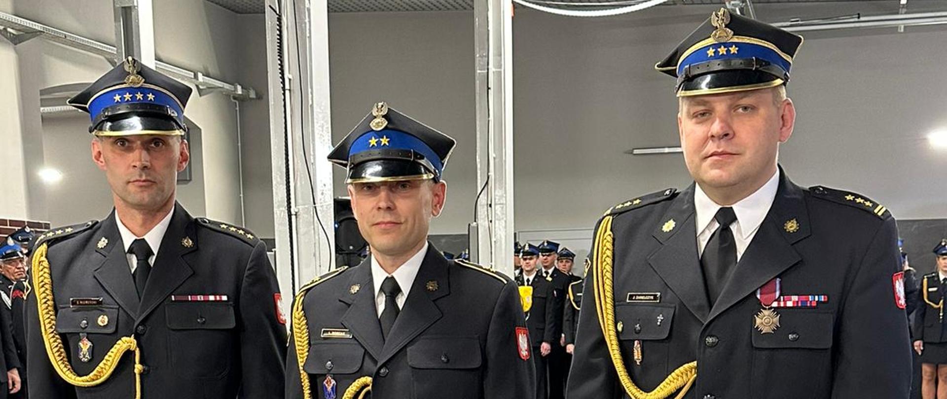 trzech strażaków w rogatywkach stoi obok siebie, w tle grupa ludzi