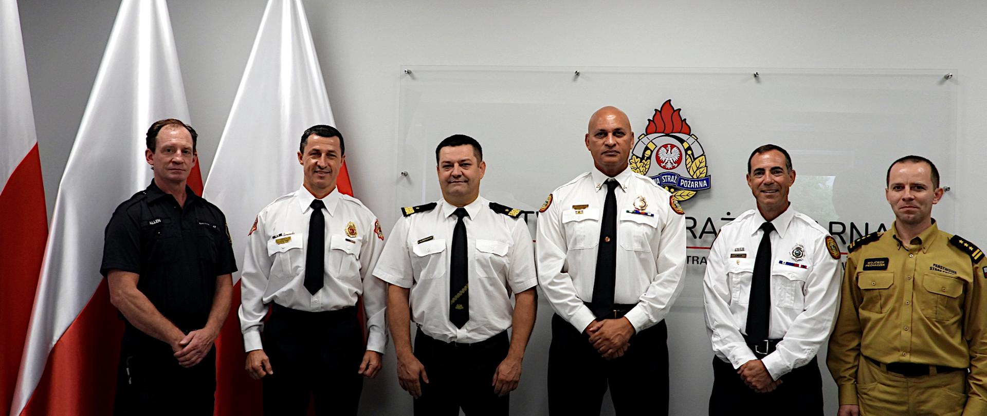 Na sali sztabowej w szeregu stoi delegacja czterech amerykańskich strażaków, śląski komendant wojewódzki PSP nadbryg. Jacek Kleszczewski oraz komendant miejski PSP w Jastrzębiu -Zdroju st. bryg. Wojciech Piechaczek. W tle na ścianie widoczne logo PSP. Po lewej stronie trzy flagi państwowe.