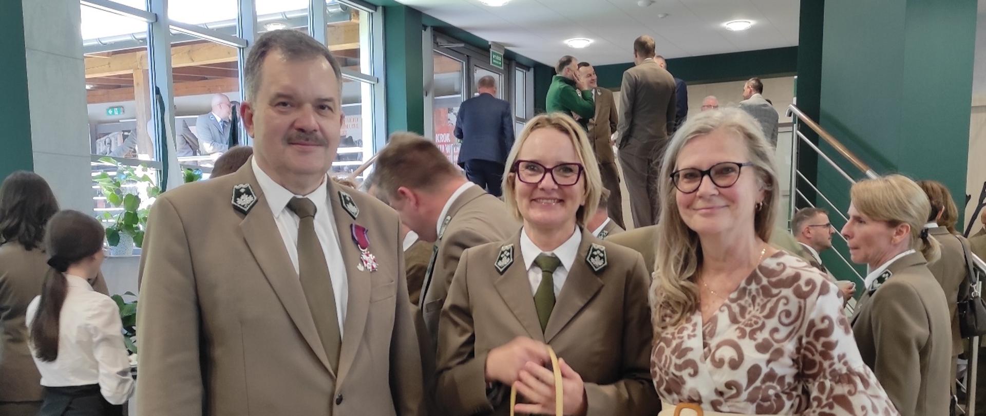 Trzy osoby pozują do zdjęcia podczas uroczystości: dwie w mundurach i kobieta w jasnej, wzorzystej sukience. W tle znajduje się grupa uczestników wydarzenia.