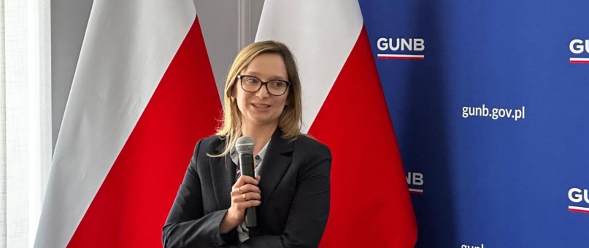 Osoba z mikrofonem na tle flag Polski i banera „GUNB” z adresem „gunb.gov.pl”, twarz zasłonięta.