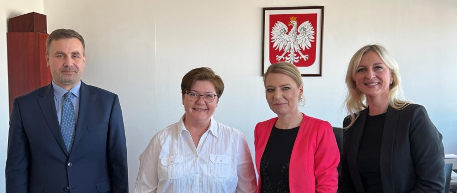 Grupa czterech osób w eleganckich ubraniach na tle ściany z godłem Polski w ramce.