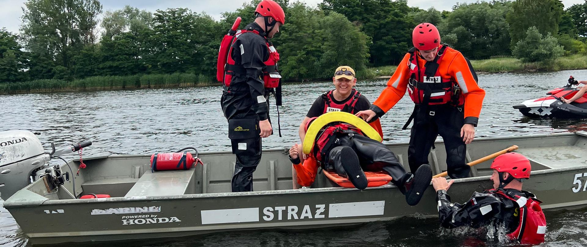 Strażacy w łodzi podczas ćwiczeń z ratownictwa wodnego pomagają wyciągnąć poszkodowaną osobę z wody na noszach